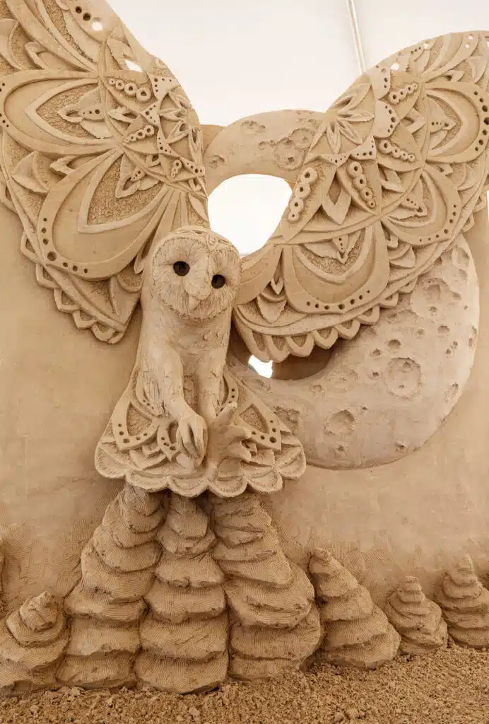 Best Things to Do in Rhode Island This Summer. Intricate sand sculpture of an owl with ornate wings, perched in front of a large moon and surrounded by cone-shaped sand formations.