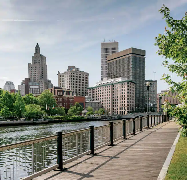 A wooden boardwalk runs beside a river, with modern and historic buildings in the background under a partly cloudy sky.