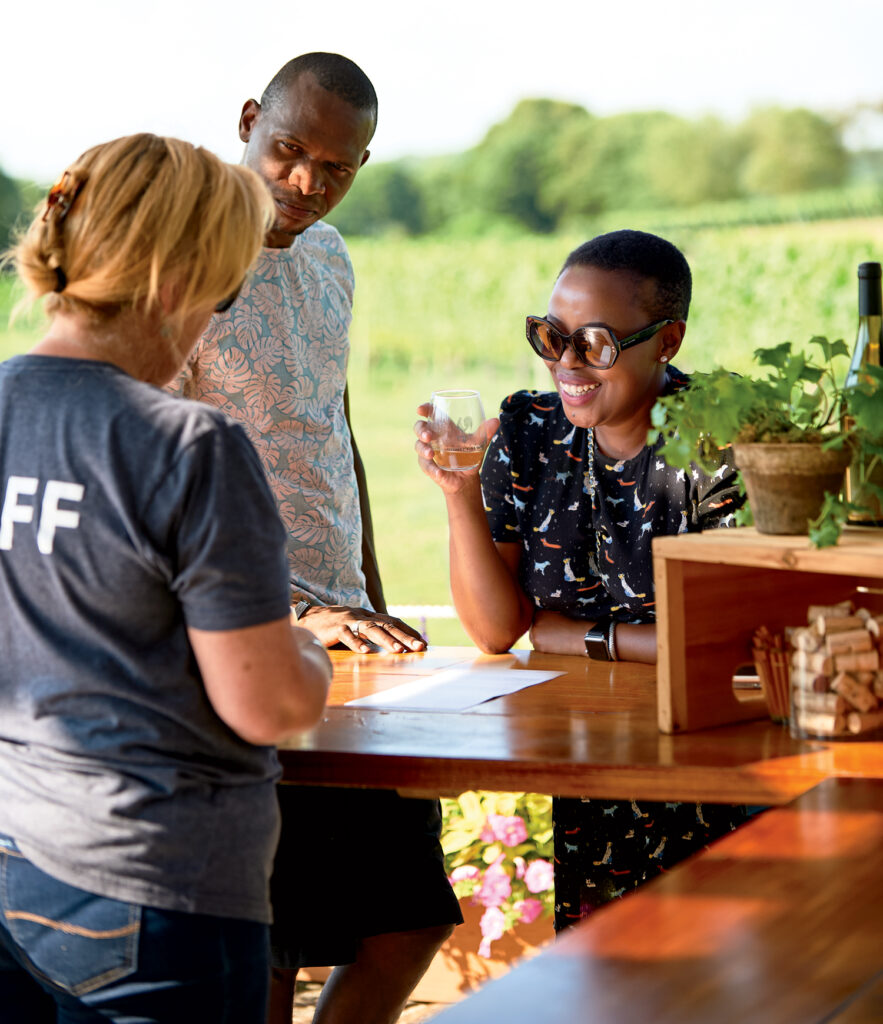 Two people stand at an outdoor wine tasting counter, chatting with a staff member during their Rhode Island travel. One person holds a glass and smiles, while wine bottles and a potted plant decorate the counter.