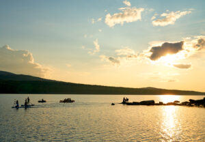 People paddleboarding, kayaking, and standing on rocks by the lake during sunset, with hills in the background and scattered clouds in the sky.