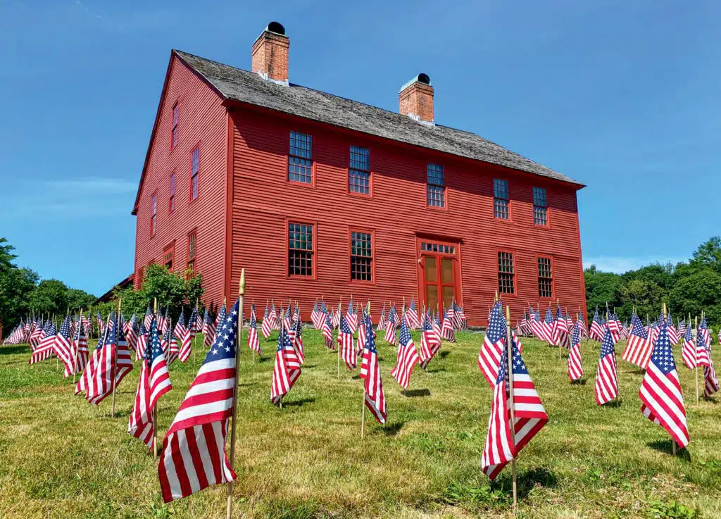 50 Must-See American Revolution Historic Sites in New England: A historic red wooden building with two chimneys stands behind a lawn filled with numerous American flags on a sunny day.
