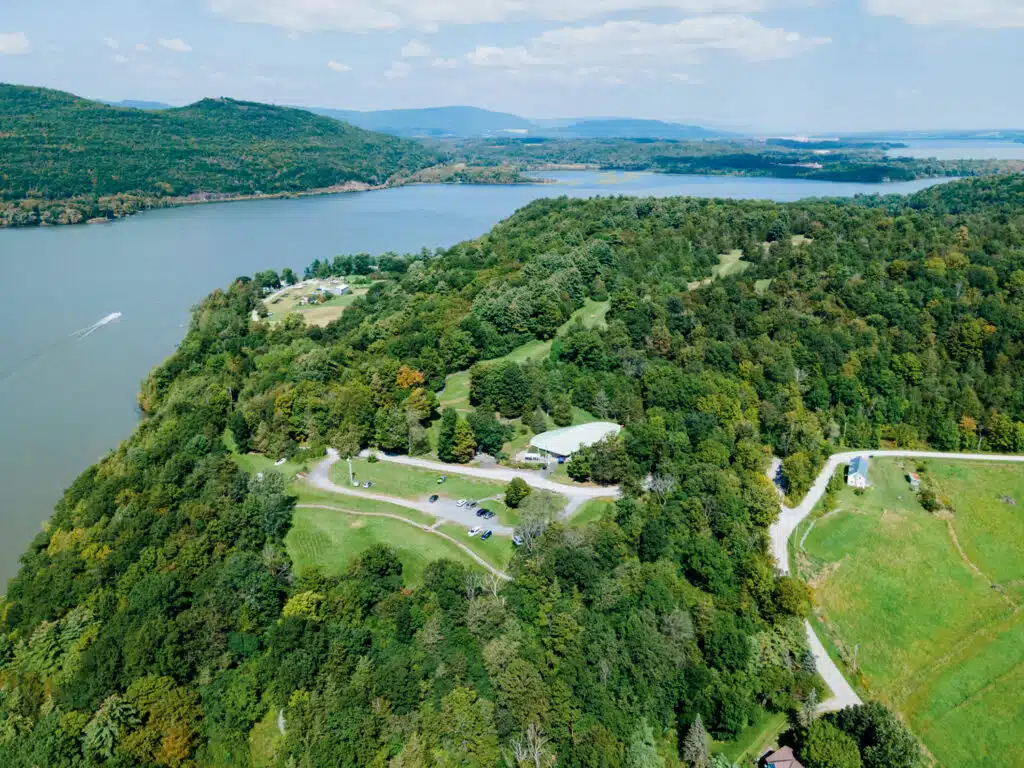 Aerial view of a hilly, forested landscape by a river, with several buildings, parking lots, grassy areas, and distant mountains under a partly cloudy sky.