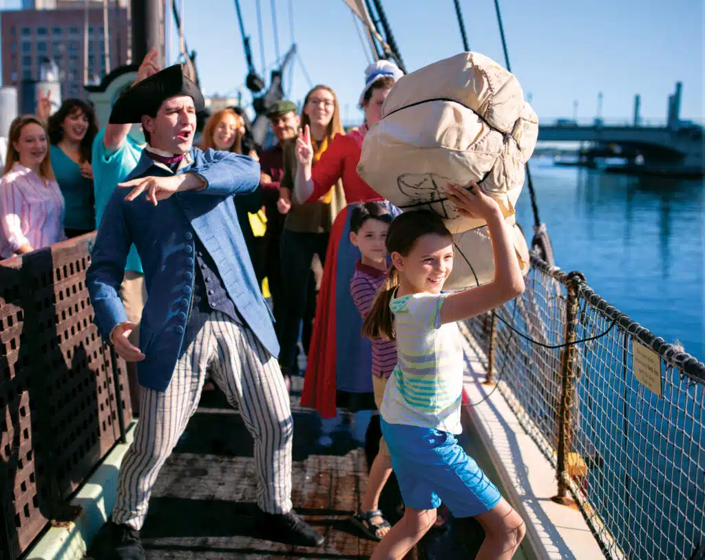50 Must-See American Revolution Historic Sites in New England: A group of people stand on a ship deck, with a child holding a large burlap sack overhead and an adult in historical clothing gesturing beside her. It is sunny and the water is visible.