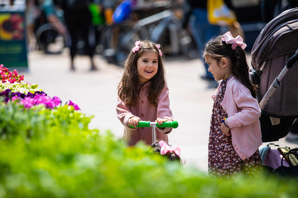 Two young girls stand near a stroller and colorful flowers outdoors; one girl holds a green scooter while both wear pink cardigans and hair bows.