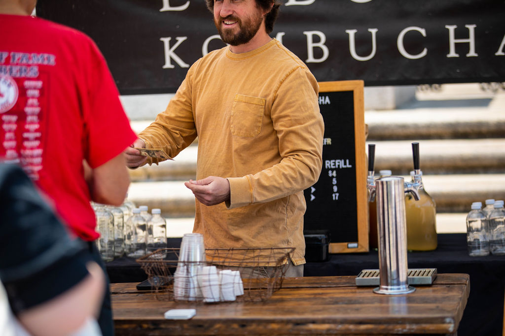A man in a tan long-sleeve shirt serves a customer at an outdoor kombucha stand with bottles and cups on display.