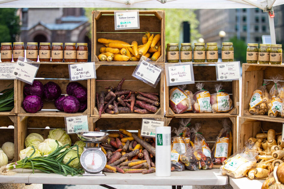 Farmers market stall displaying jars of preserves, red and yellow carrots, parsnips, red cabbage, green onions, a scale, and packaged goods arranged in wooden crates.