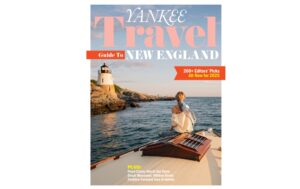 A woman sits on a boat facing a coastal lighthouse at sunset, featured on the cover of Yankee Travel Guide to New England.