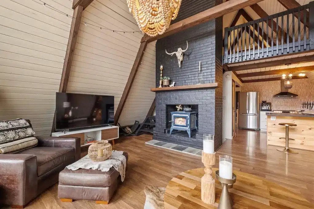 A-frame cabin living area with wood floors, a black brick fireplace, mounted bull skull, leather chair, TV, and open kitchen with loft above.