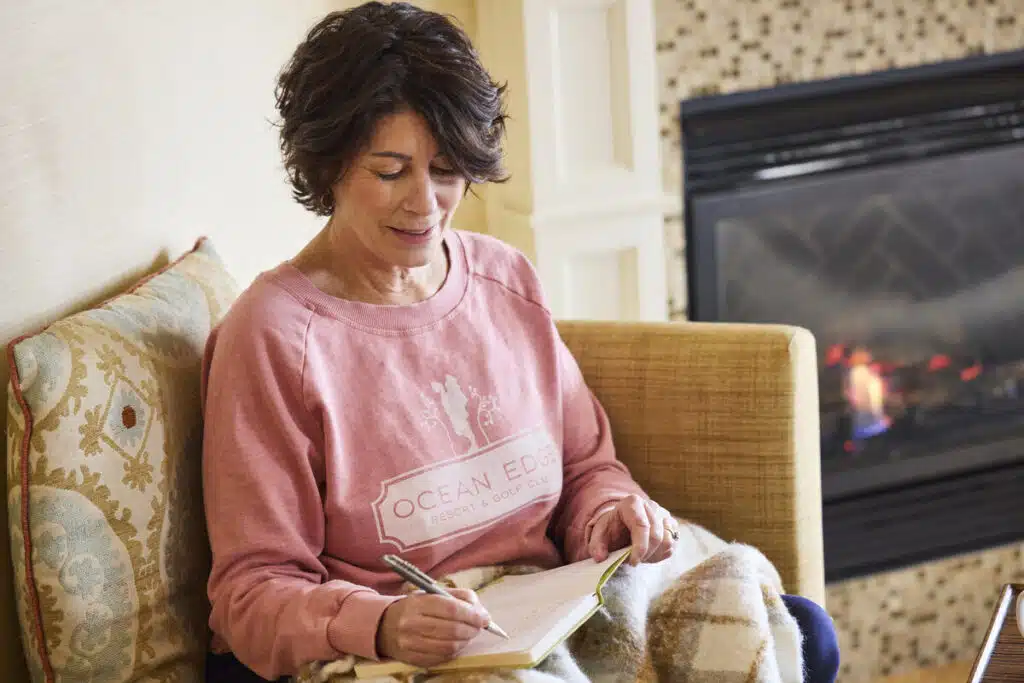 Woman in a pink sweatshirt writes in a notebook while sitting by a fireplace, covered with a blanket.