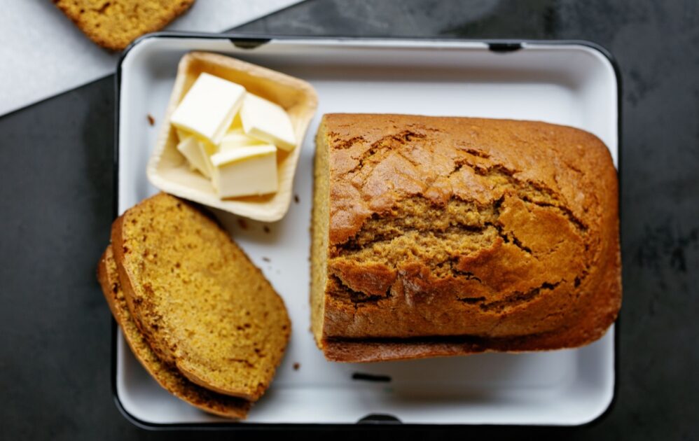 A loaf of pumpkin bread with two slices cut, served on a white tray next to a small dish of butter.