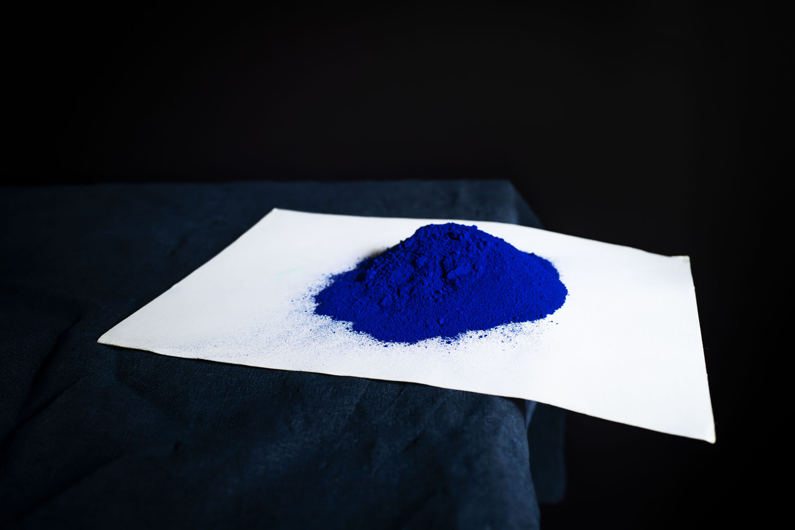A pile of bright blue powder sits on a white sheet of paper, positioned on a dark fabric surface with a black background.