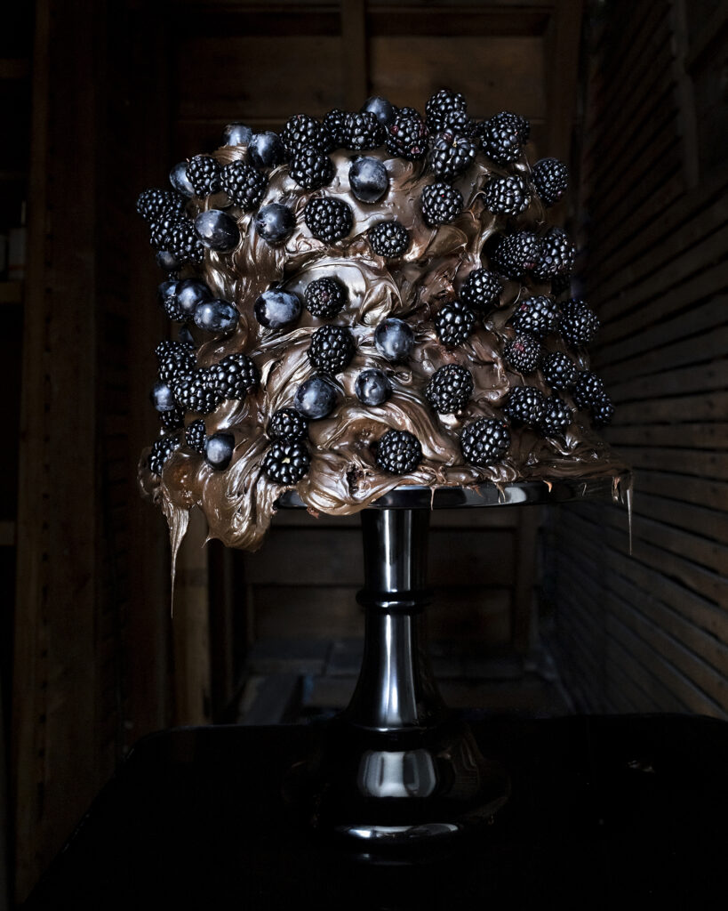 A tall cake with metallic gold icing, topped and covered with blackberries and dark berries, displayed on a black pedestal stand.