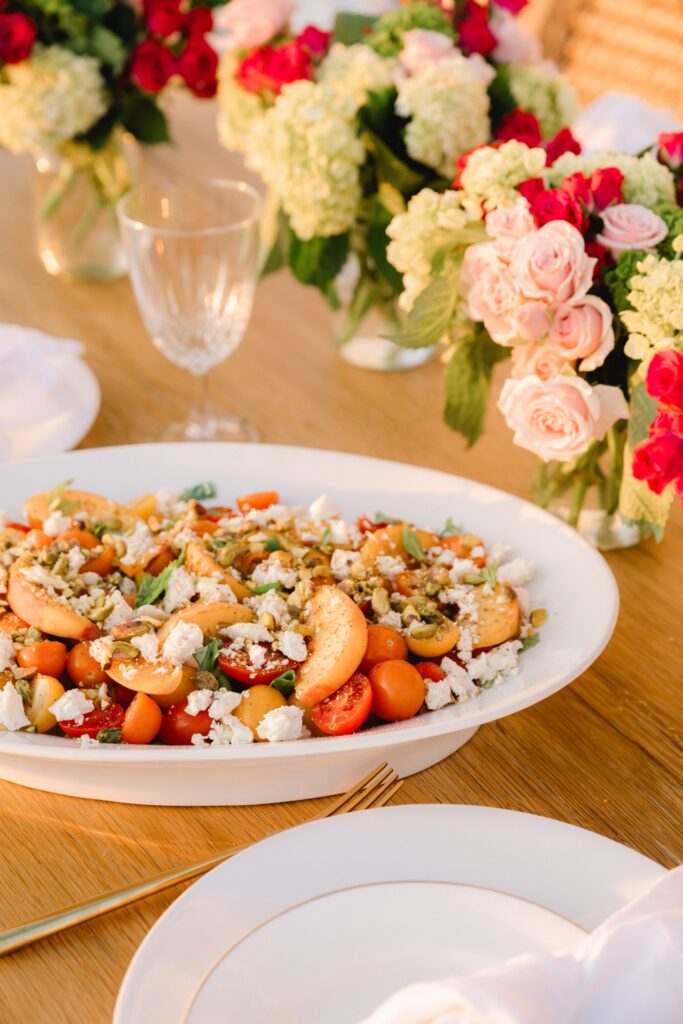 A white plate of salad with peaches, cherry tomatoes, feta cheese, and herbs sits on a wooden table set with glassware and floral centerpieces.