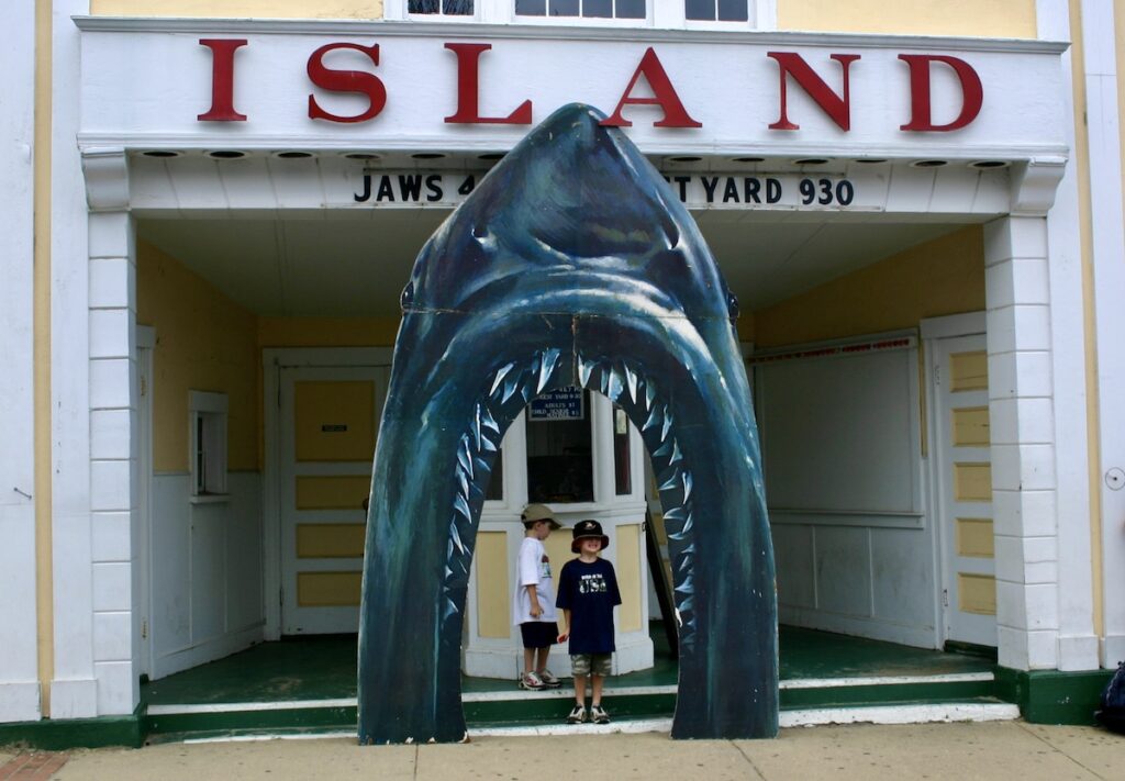 Two children stand inside a large shark jaw display at the entrance of a building labeled "ISLAND" and "JAWS COURTYARD 930.