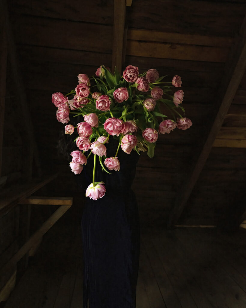 A person stands indoors holding a large bouquet of pink tulips that obscures their face and upper body; wooden beams are visible in the background.