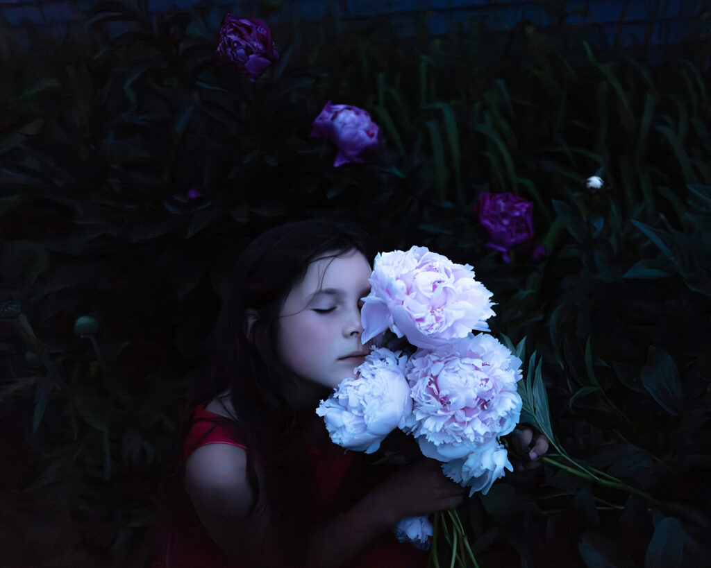 A girl in a red dress holds and smells a bouquet of pale pink peonies in a dark garden with purple flowers in the background.