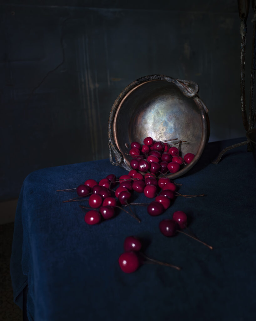 A tarnished metal bowl tipped over on a dark blue tablecloth, spilling a cluster of ripe red cherries across the surface.