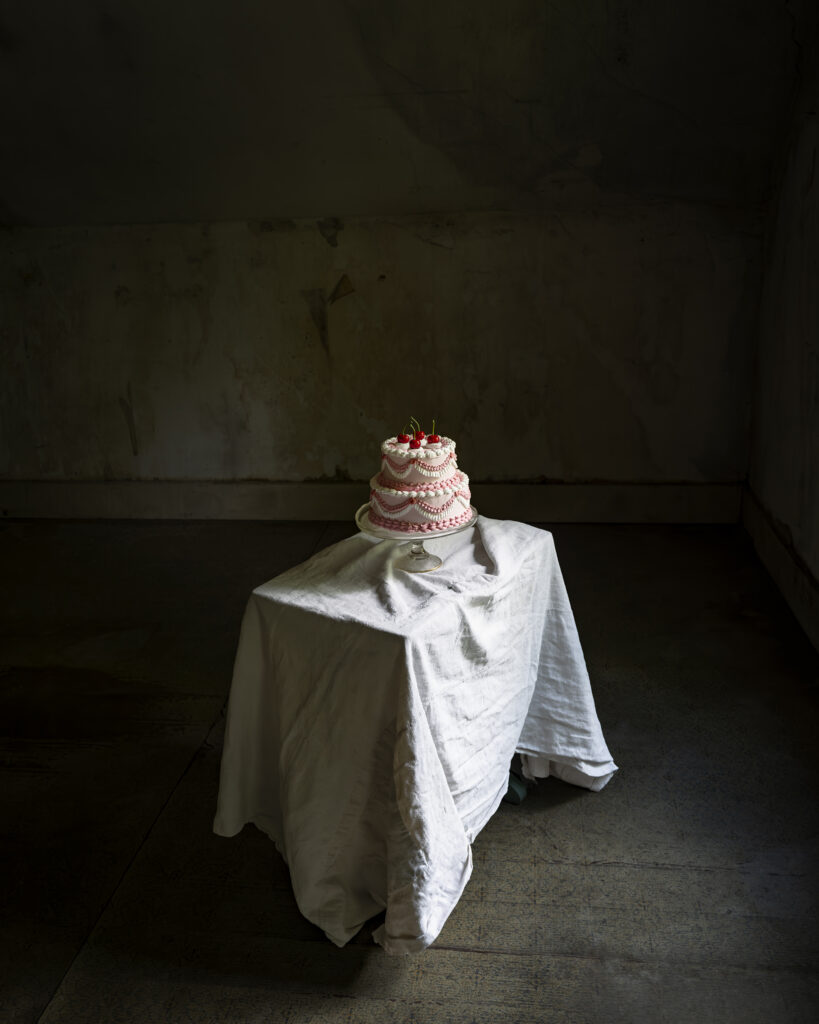 A three-tiered pink and white cake with strawberries on top sits on a small table covered with a white cloth in a dimly lit, empty room.