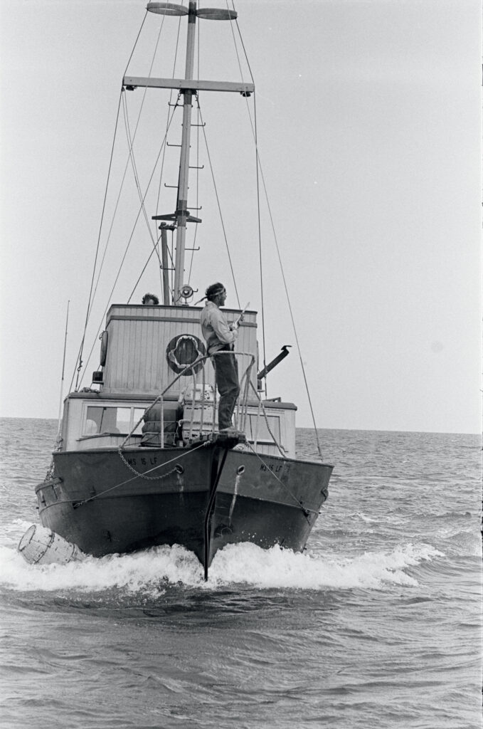 A person stands at the bow of a small boat moving through calm ocean water, with the front of the vessel creating a wake.