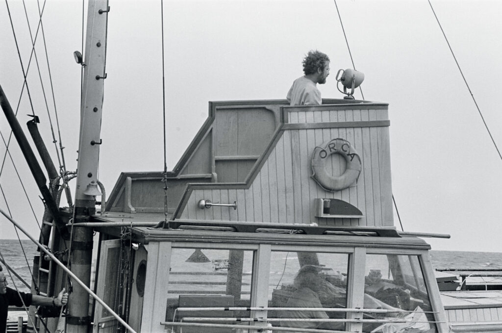 A person stands on the upper deck of a boat named "Orca," holding a loudspeaker, while two others sit below; the ocean and a dorsal fin are visible in the background.