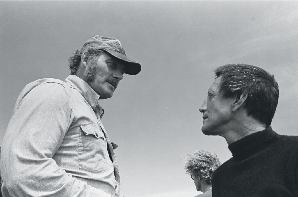 Two men face each other outdoors, one wearing a cap and denim shirt, the other in a dark sweater; a third person is partially visible in the background under a clear sky.