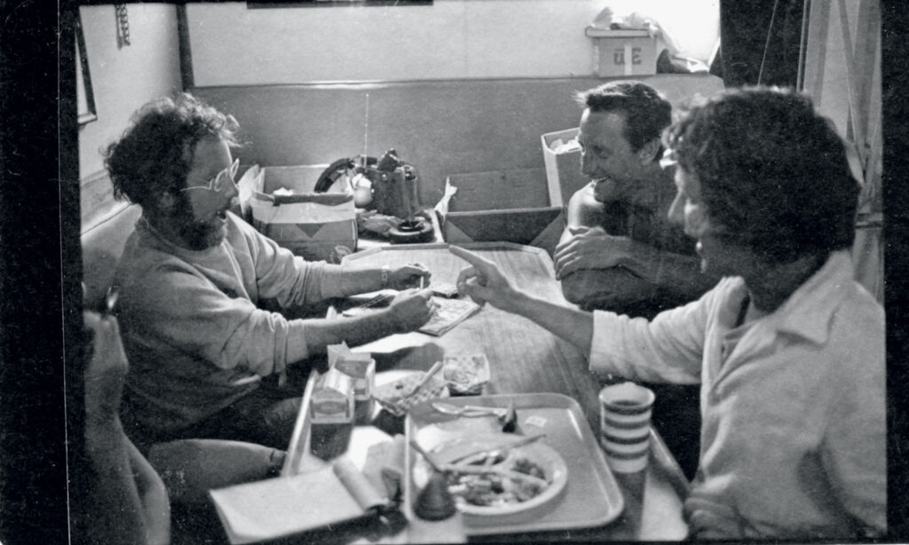 Four people sit around a table playing cards and talking. Food trays, drinks, and various items are on the table in a casual indoor setting.