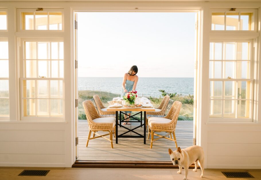 Debi Lilly sets an outdoor dining table on a deck overlooking the ocean, while a small dog stands inside near the open doors.