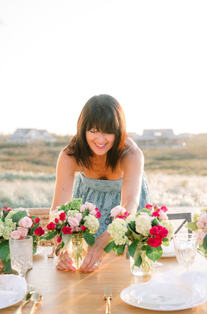 A woman arranges floral centerpieces on an outdoor dining table set with plates and glassware, with a bright, natural background.