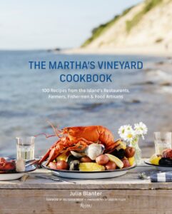 A cookbook titled "The Martha’s Vineyard Cookbook" sits on a table by the beach with a plate of lobster, corn, and potatoes in the foreground.