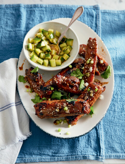 Matt’s Favorite Finger Lickin’ Good Sweet and Sour Ribs. A white oval plate with glazed ribs topped with sesame seeds and chopped green onions, garnished with cilantro, next to a small bowl of diced cucumber salad.