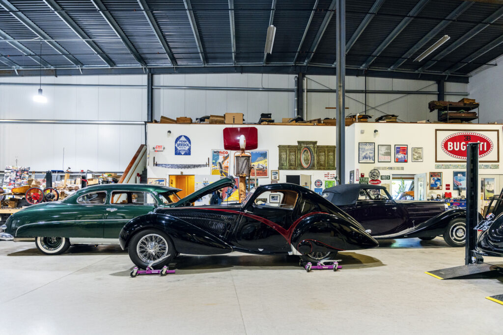 Three vintage cars are displayed side by side inside a spacious, well-lit garage with automotive memorabilia on the walls and shelves in the background.