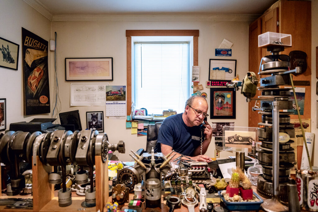 Meet Vermont Bugatti Restoration Expert Scott Sargent. A man sits at a cluttered desk filled with mechanical parts and papers, talking on the phone in a room decorated with car posters and framed pictures.
