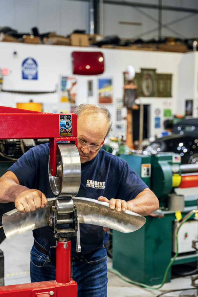 A man operates a metalworking machine in a workshop, shaping a curved sheet of metal with both hands.