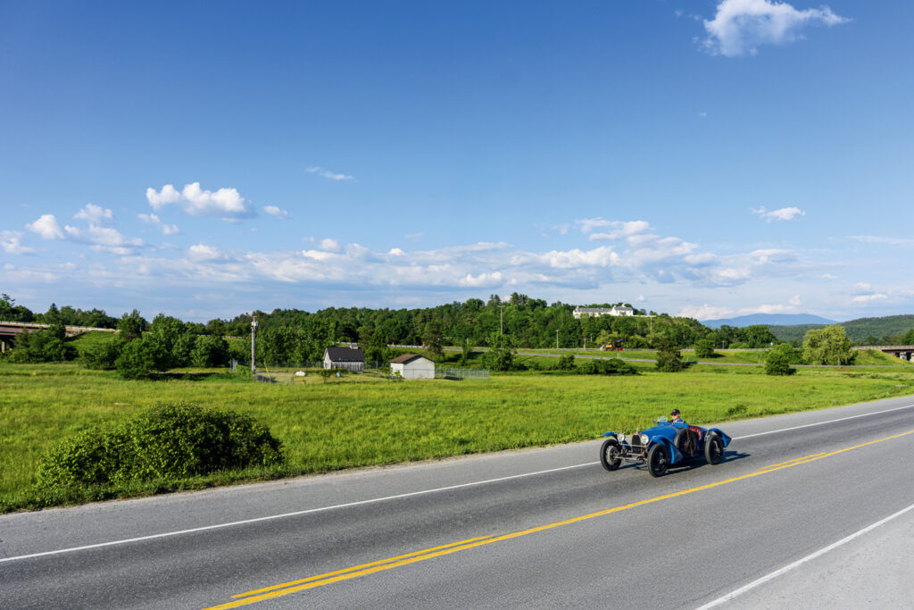 A person drives a vintage blue car on a rural road, with green fields and scattered buildings under a blue sky with scattered clouds.
