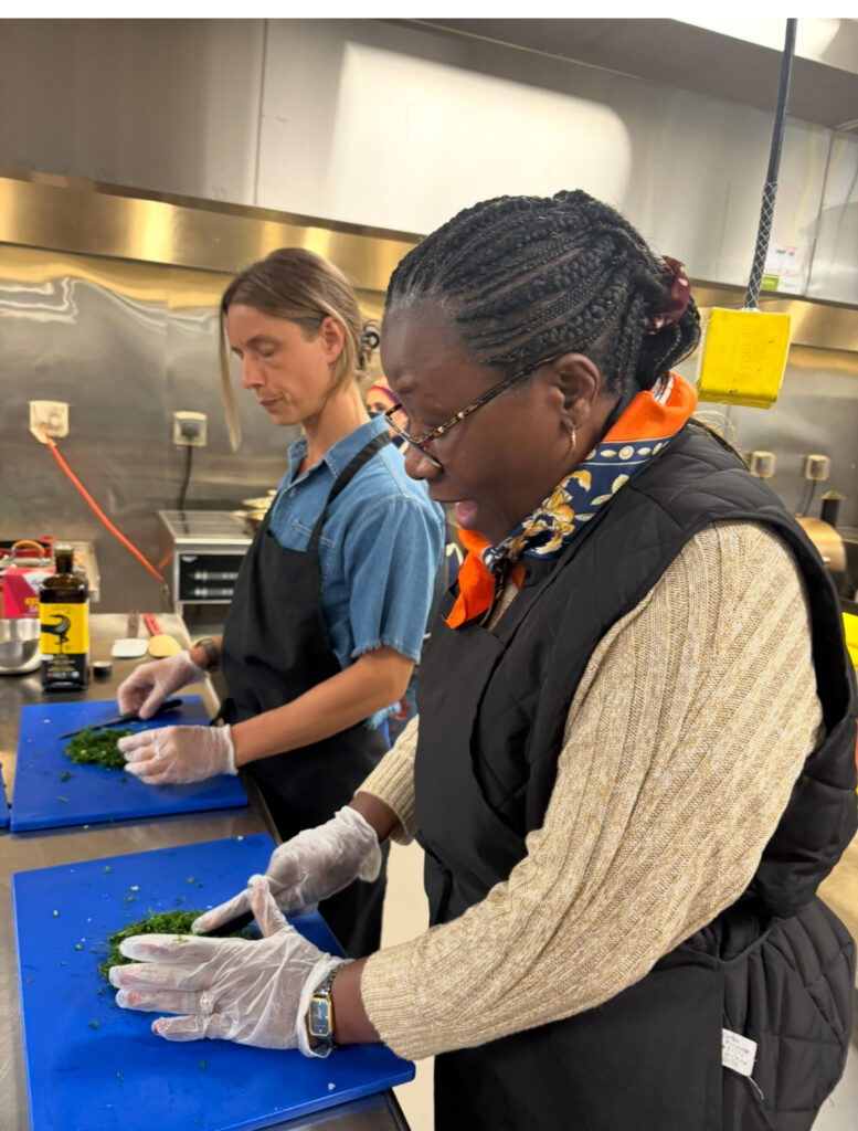 Two people wearing aprons and gloves chop herbs on blue cutting boards in a commercial kitchen.