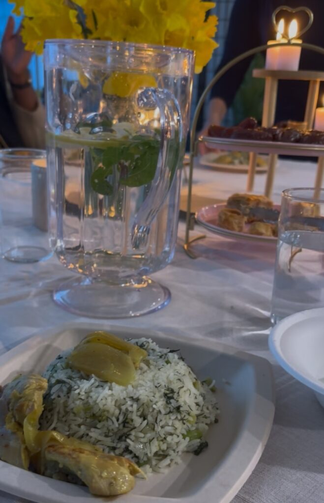 A plate of rice with artichokes sits on a white tablecloth beside a glass pitcher of water with mint and lemon, flowers, and a tiered tray of snacks.