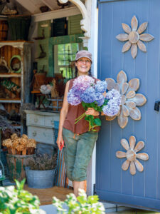 Hydrangea Care and Pruning Tips for Big, Beautiful Blooms. A woman holding a bouquet of hydrangeas stands in the doorway of a garden shed decorated with large wooden flowers.