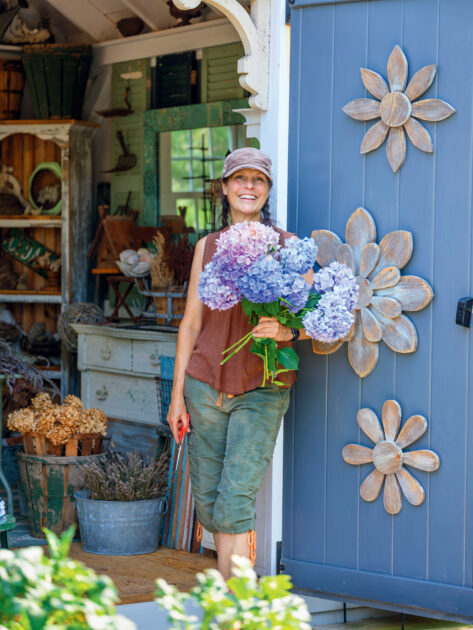 Hydrangea Care and Pruning Tips for Big, Beautiful Blooms. A woman holding a bouquet of hydrangeas stands in the doorway of a garden shed decorated with large wooden flowers.