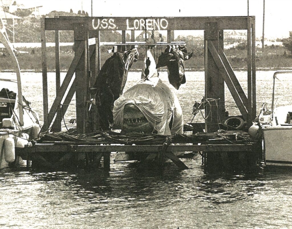 Black-and-white photo of a wooden floating dock labeled "U.S.S. LORENO" with equipment and covered items on it, moored between two boats on the water.