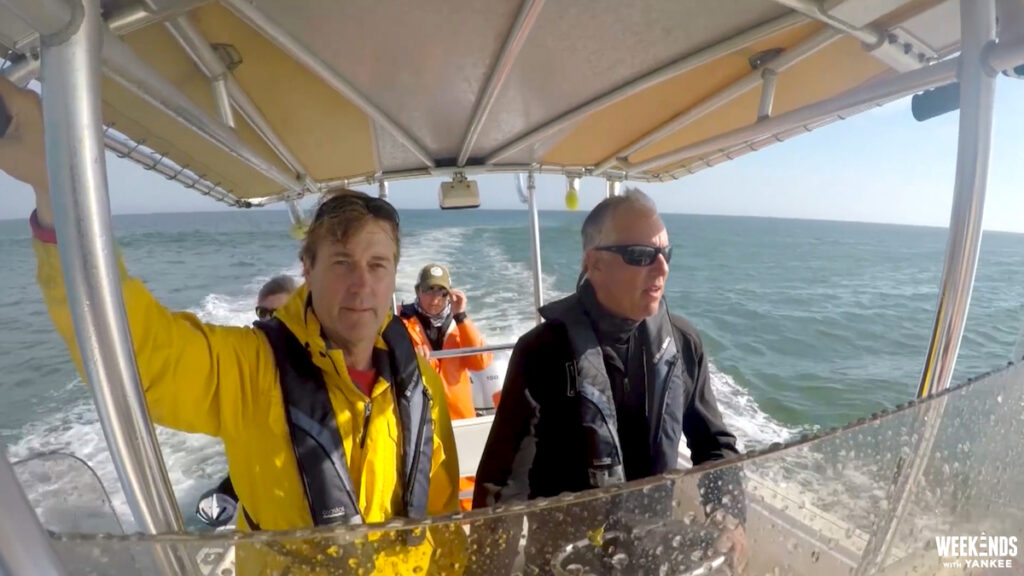 Four people wearing life jackets ride on a small motorboat across the ocean under a sunny sky, with two people visible at the front and waves in the background.