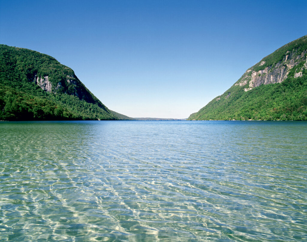 Clear blue water with gentle ripples stretches between two green, tree-covered hills under a bright blue sky.