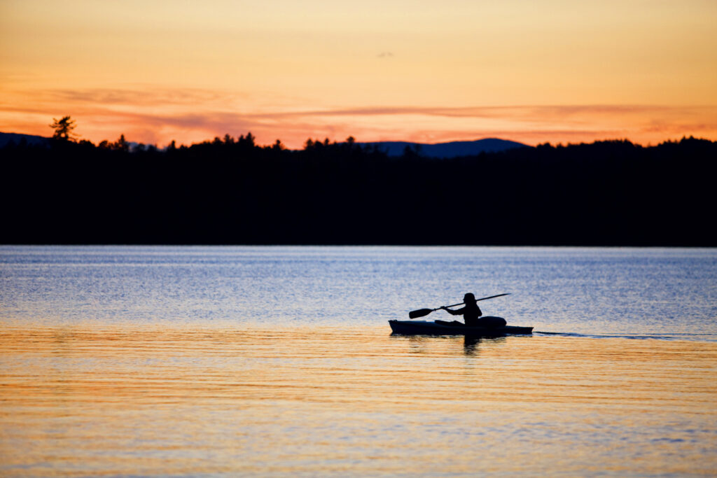 Lakeside Activities and Attractions in New England. A person kayaking on a calm lake at sunset, with trees and mountains visible in the background.
