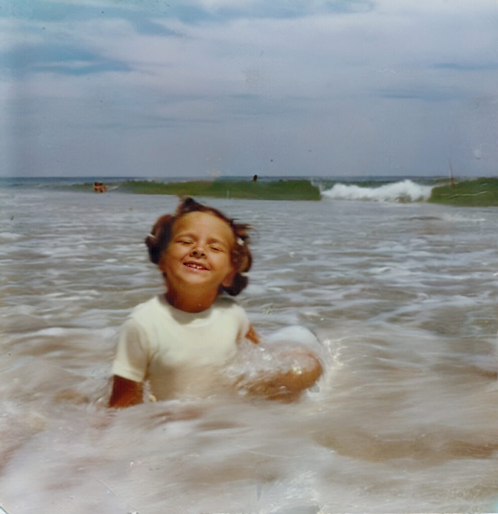 The Great Ocean vs. Lake Debate. Young child with short brown hair smiles while sitting in shallow ocean waves on a beach, wearing a white shirt, with blue sky and distant horizon visible in the background.