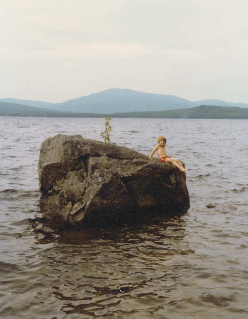 A child in red swimwear sits on a large rock in a lake, with a small plant growing on the rock and mountains in the background.