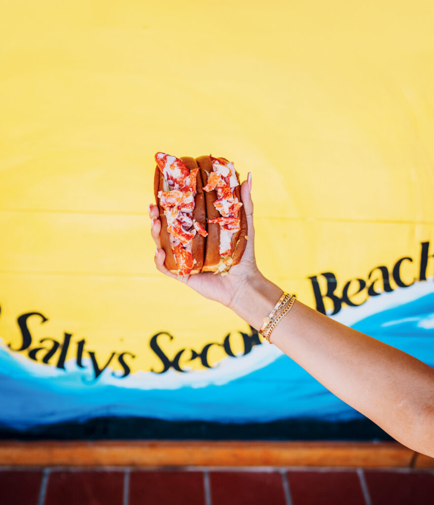Best Waterfront Restaurants in New England: Coastal and Lakefront Dining Picks. A hand holding a lobster roll in front of a yellow and blue mural with partially visible text.