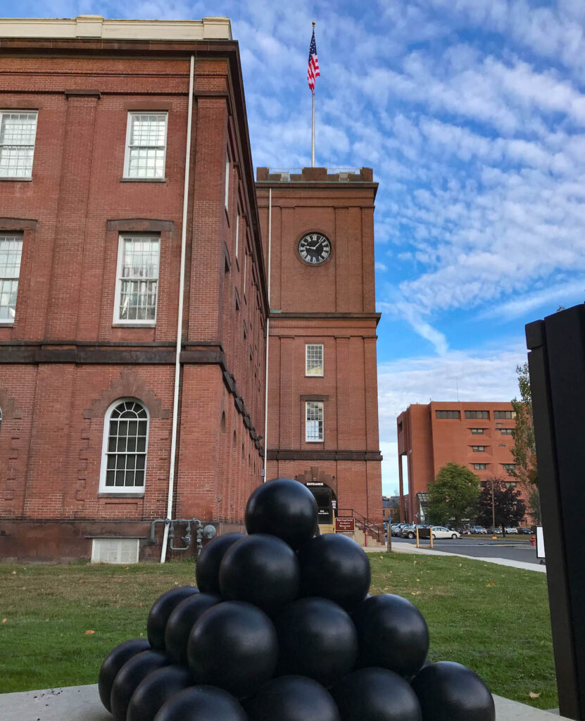 A stack of black cannonballs sits on grass in front of a red brick building with a clock tower and a U.S. flag on top.