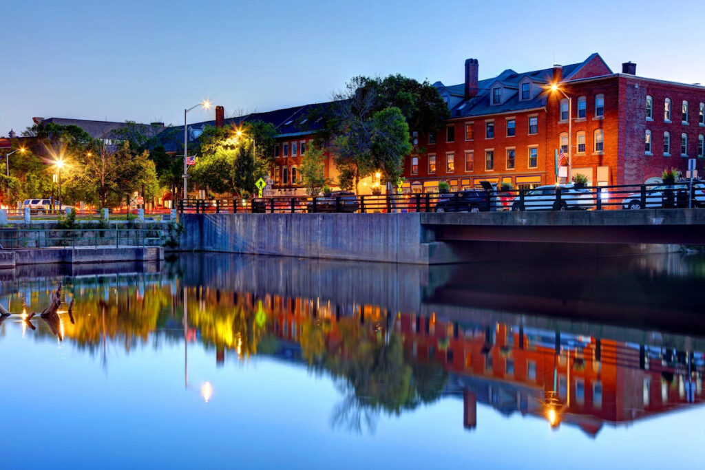 A calm river reflects brick buildings, trees, and streetlights at dusk, with a bridge crossing over the water in an urban setting.