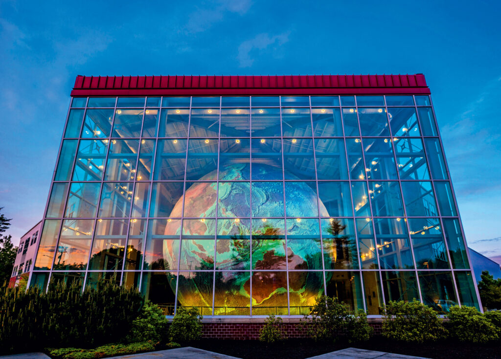 A large globe is displayed inside a glass-walled building at sunset, with reflections of lights and the sky visible on the structure.