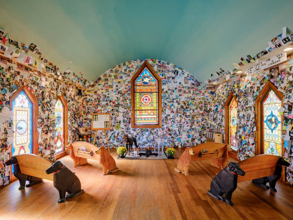 A small chapel interior features stained glass windows, walls covered with photographs, and pews shaped like dogs. Four carved dog figures sit near the center.