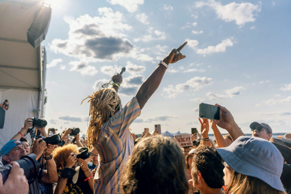 A performer with blond dreadlocks holds a microphone and points skyward on an outdoor stage, surrounded by a crowd taking photos under a partly cloudy sky.